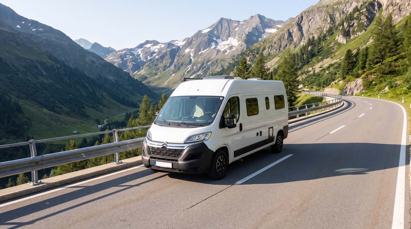 Weißer Kastenwagen-Camper mit festem Hochdach auf einer breiten Passstraße in den Alpen, links eine Leitplanke über dem Tal, dahinter schroffe Felsgipfel mit Restschnee im klaren Morgenlicht