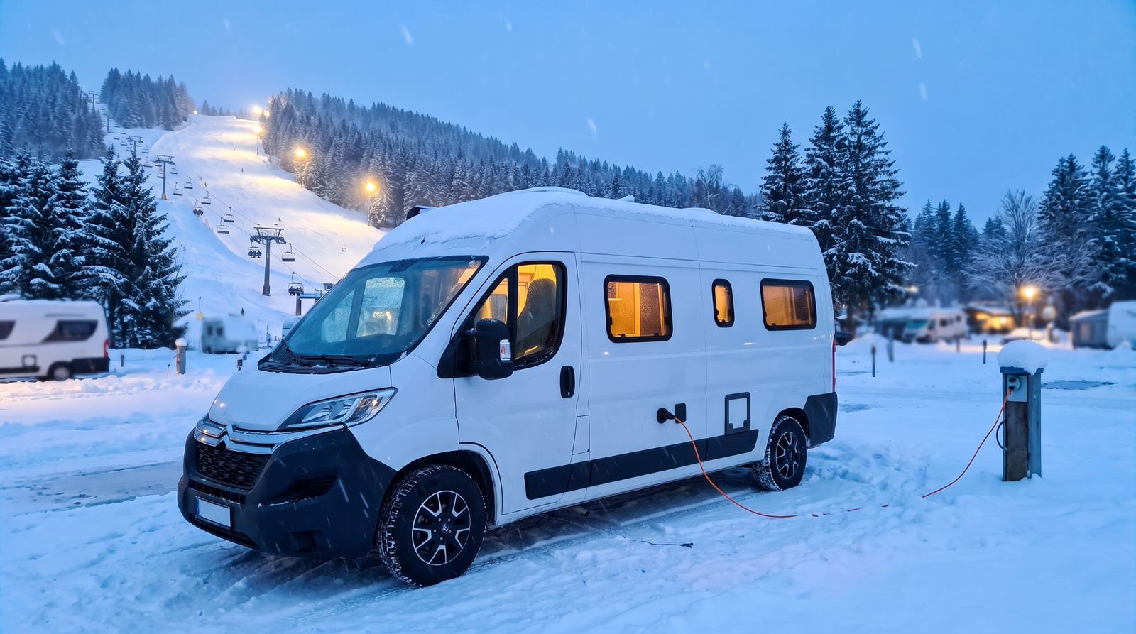 Weißer Kastenwagen-Camper mit festem Hochdach auf einem verschneiten Campingplatz zur blauen Stunde, Stromkabel im Schnee, im Hintergrund eine beleuchtete Skipiste mit Sessellift