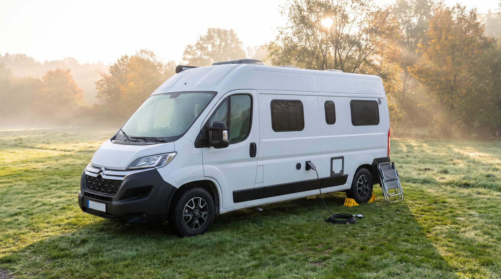 Weißer Kastenwagen-Camper auf einer Wiese im Morgennebel, das Stromkabel steckt noch, gelbe Auffahrkeile liegen am Hinterrad und ein zusammengeklappter Campingstuhl lehnt daneben
